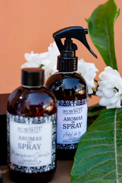 Botellas de aroma en spray de la línea Ámbar, una de color oscuro con etiqueta de "Aromas en Spray" y otra de "Aromas Spray - Manzana Canela", rodeadas de flores blancas y hojas verdes.