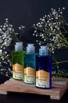 Botellas de espumas de baño mini aromARTE en colores verde, azul y amarillo, presentadas sobre una base de madera con flores blancas al fondo. aromas jazmín, manzana y lavanda