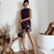 Mulher vestindo vestido curto preto com gola alta, estampa de borboletas e flores na barra, segurando bolsa preta, em ambiente decorado com plantas secas e vaso branco.
