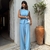 Mulher vestindo conjunto azul feminino composto por blusa sem mangas e calça pantalona com cinto, em ambiente externo com vaso de planta ao fundo.