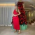 Mulher vestindo um vestido midi vermelho de tricô modal um ombro, posando ao lado de uma árvore de Natal decorada com enfeites vermelhos.