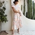 Mulher vestindo vestido midi floral com cinto Ana em ambiente externo decorado com plantas, ideal para ocasiões elegantes e casuais.