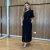 Mulher vestindo conjunto feminino composto por blusa de um ombro só e saia midi preta, posando em ambiente interno com parede de madeira ao fundo.