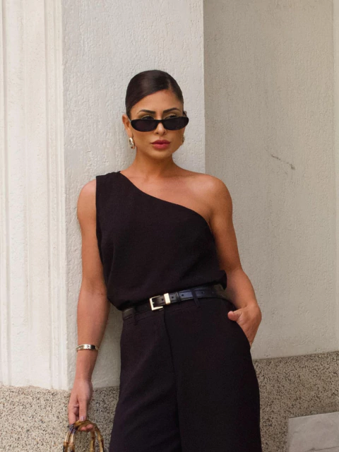Mulher usando blusa preta de ombro único com calça preta e cinto, segurando bolsa com alça de bambu