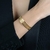 Bracelete dourado de renda vazado em um pulso feminino, destacando seu design elegante e moderno. Tenure Semijoias