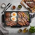 Parrilla grill eléctrica antiadherente SUONO de 49x27cm cocinando hamburguesas, huevos, salchichas, vegetales y bacon en una mesa con especias y utensilios de cocina.