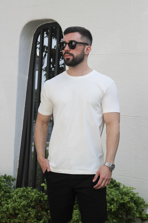 Homem vestindo uma camiseta básica off white, com óculos escuros e calças escuras, em um ambiente ao ar livre.