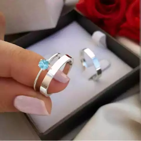 Par de alianças de namoro em prata 950, uma com pedra azul e outra lisa, sendo seguradas por uma mão feminina com unhas pintadas de rosa claro, caixa de joias ao fundo