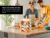 Especiero de bambú natural con 12 tarros de vidrio para especias sobre una mesada de cocina, con dos personas cocinando al fondo