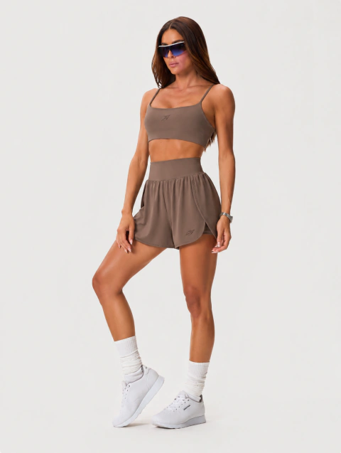 Mulher usando shorts feminino marrom Cristal da marca Mormaii, combinado com top esportivo e tênis branco