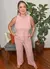 Mulher usando um conjunto rosa listrado, posando com as mãos nos bolsos, em um ambiente interno com plantas ao fundo.