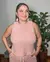 Mulher usando um conjunto rosa listrado, posando com as mãos nos bolsos, em um ambiente interno com plantas ao fundo.