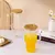 Jarro de vidrio con tapa de bambú y sorbete, relleno con bebida naranja y cubitos de hielo, sobre una mesa blanca con otro jarro vacío y elementos decorativos