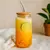 Vaso de vidrio con tapa de bambú y sorbete de vidrio, lleno con bebida refrescante naranja con rodaja de lima y perlas en el fondo