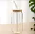 Vaso de vidrio transparente con tapa de bambú y sorbete de vidrio sobre mesa de madera clara