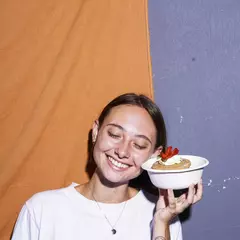 Mujer sonriente sosteniendo un bowl blanco con un postre decorado con crema y fresas, frente a un fondo colorido.