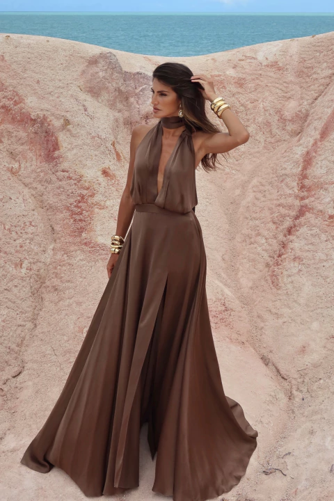 Mulher usando vestido de seda marrom Layla Luminare com decote profundo e saia ampla, posando em cenário natural com fundo de falésias e mar ao fundo.