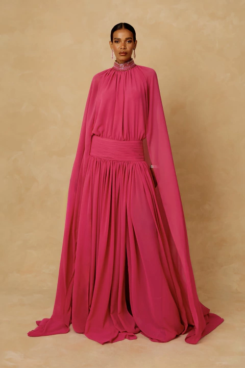 Modelo vestindo vestido longo Áurea Bordado na cor pink, com gola alta bordada em pedrarias, capa integrada e saia fluida com drapeado na cintura.