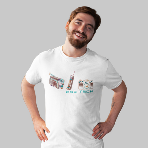Camiseta branca com estampa retrô de ícones tecnológicos dos anos 80: filmadora VHS, câmera fotográfica analógica e telefone sem fio. Produto da marca Nas Antigas, modelado por homem sorridente em fundo neutro.
