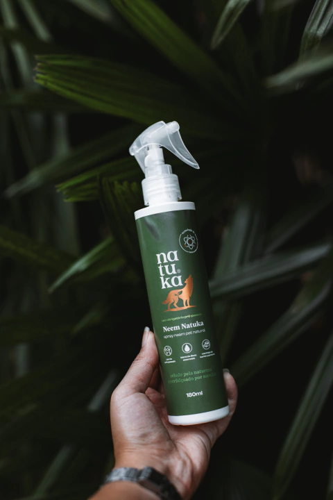 Embalagem de Natuka Spray NEEM de 180 ml, lata de cor verde escuro com detalhes em branco e laranja em um plano de fundo com plantas verde escuro.