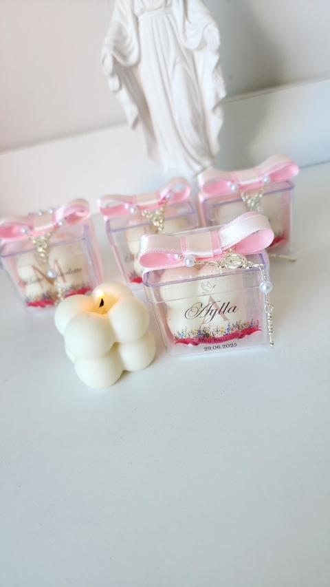 Kit com 10 lembrancinhas de batizado com velas bubble brancas em embalagem transparente decorada com laço rosa e terço, ao fundo está uma estátua religiosa branca.