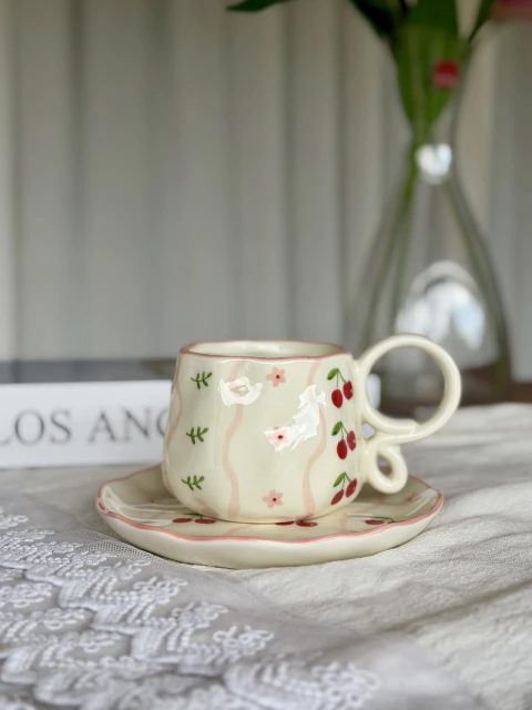 Taza de cerámica con diseño de cerezas y flores pequeñas sobre plato a juego, ambos con borde rosado