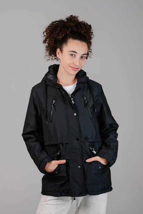 Modelo femenina con campera negra Islandia, capucha fija, bolsillos con cierres y detalles ajustables, posando con manos en los bolsillos