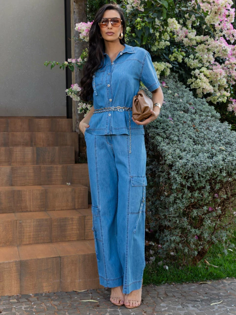 Mulher vestindo conjunto denim com pesponto duplo, composto por camisa de manga curta e calça jeans de modelagem ampla, em ambiente externo com flores ao fundo.