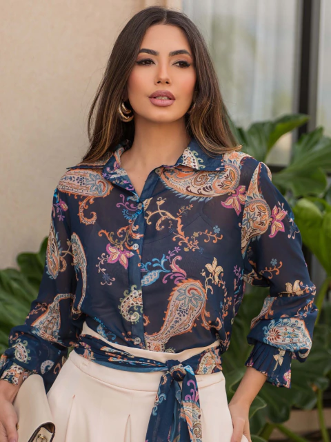 Mulher vestindo camisa chiffon estampada com detalhe plissado, padrão colorido em fundo azul escuro, combinada com saia clara. Moda feminina elegante.