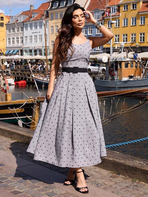 Mulher usando vestido tricoline fio tinto vichy com pregas e estampa de poá, cinto preto na cintura e sandálias pretas, em cenário urbano à beira de um canal com barcos e casas coloridas ao fundo.