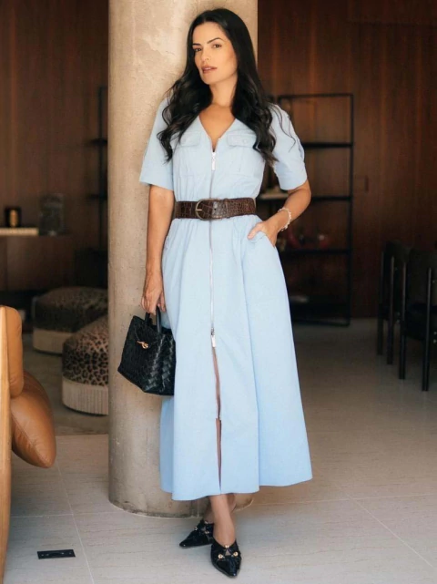 Mulher vestindo vestido chemisier de sarja azul claro com bolsos utilitários, cinto marrom e bolsa preta, em ambiente interno moderno.