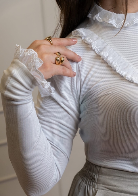 Blusa branca de manga comprida com detalhes em babado de renda na gola e nos punhos, usada por modelo com anéis dourados