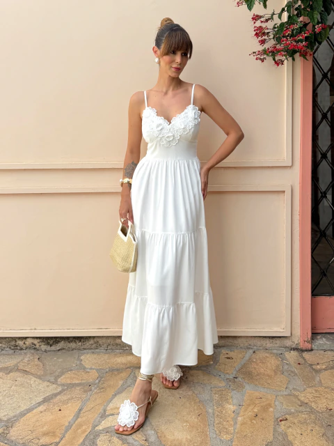Mulher usando vestido branco longo com alças finas e detalhes florais no busto, segurando bolsa de palha