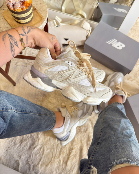 Tenis New Balance 9060 bege em destaque, com detalhes em branco e cinza, sendo segurado por uma pessoa com tatuagens, enquanto usa jeans.