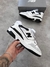 Tênis 550 branco com detalhes em preto, modelo premium, sendo segurado por uma mão masculina com pulseira e tatuagem, caixa cinza ao fundo