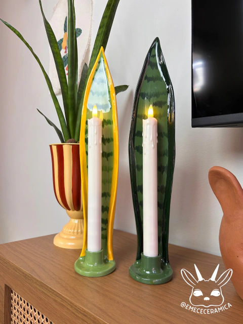 Porta vela em cerâmica em formato de folhas verdes com borda amarela e verde escuro, contendo velas brancas acesas, sobre móvel de madeira com vaso e decoração ao fundo