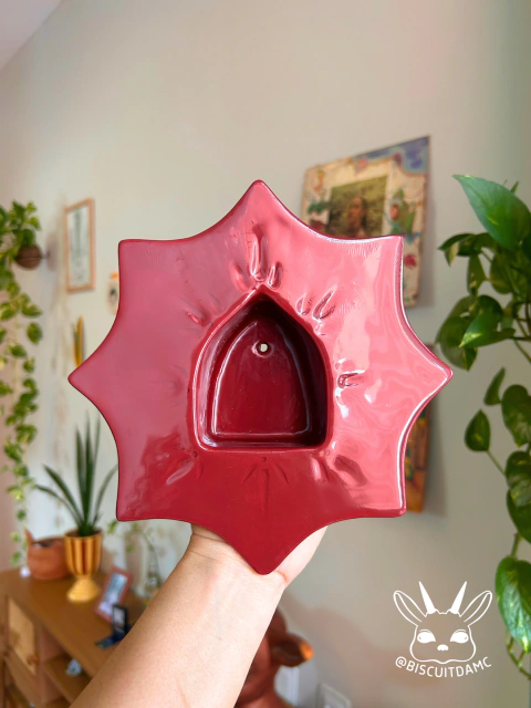 Porta-vela vermelho em formato de estrela em cerâmica fria com cavidade para vela, sendo segurado por uma mão em ambiente interno com plantas ao fundo