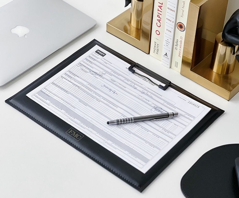 Prancheta horizontal em couro sintético preto personalizada sobre uma mesa de escritório, com um formulário preso e uma caneta posicionada em cima.