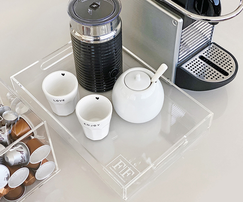 Bandeja retangular de acrílico cristal 30 x 20 cm com itens de café organizados, incluindo xícaras, pote de açúcar e espumador de leite, ao lado de uma máquina de café.
