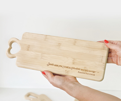 Tábua de bambu baguete personalizada com alça em formato de coração, sendo segurada por uma pessoa. Ideal para servir frios e pães.