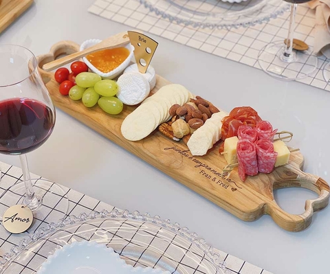 Tábua de frios retangular personalizada com alças em formato de coração, servida com queijos, salame, frutas, castanhas e biscoitos, acompanhada de uma taça de vinho tinto.