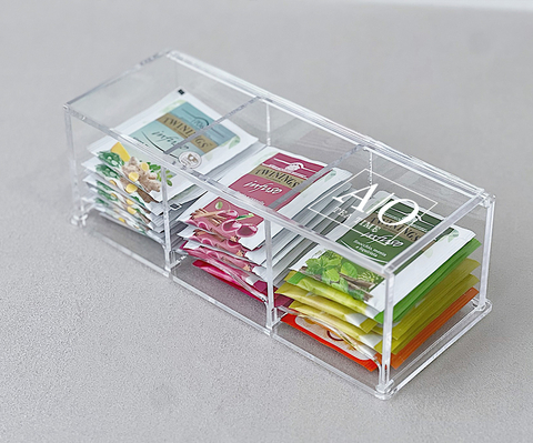 Caixa de acrílico transparente com três divisórias, contendo sachês de chá organizados por cor e sabor.