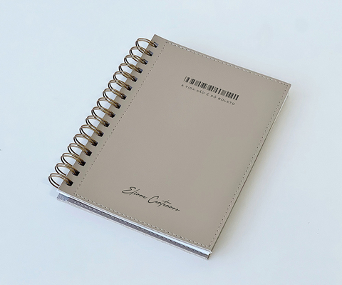 Caderno agenda permanente personalizado com capa bege, espiral metálico dourado e inscrição "A vida não é do jeito" na capa. Produto de escritório ideal para organização pessoal.