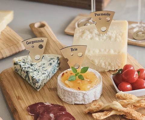 Identificadores para queijo personalizados em formato de fatia, marcando diferentes tipos de queijos em uma tábua de frios com gorgonzola, brie e parmesão.
