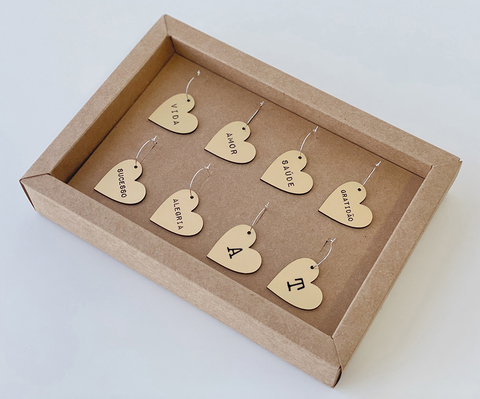 Conjunto de marcadores de taças em formato de coração personalizados, feito de material dourado, exibidos em uma caixa de papelão. Cada marcador possui uma palavra diferente gravada, ideal para identificar taças em eventos e festas.