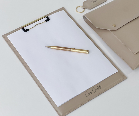 Prancheta A4 personalizada na cor bege com folha de papel e caneta dourada sobre uma mesa, acompanhada de pasta e chaveiro combinando, ideal para escritório.