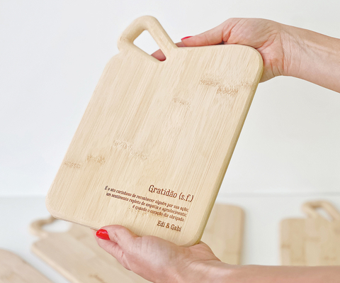Tábua de bambu retangular com cabo e gravação personalizada, sendo segurada por mãos femininas. Ideal para servir frios e queijos.