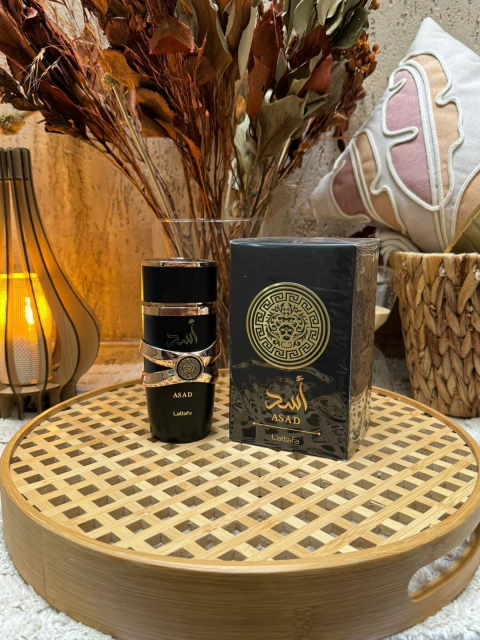 Frasco e embalagem do perfume árabe masculino Asad Lattafa Eau De Parfum 100 ml sobre bandeja de madeira com decoração de flores secas e almofada ao fundo