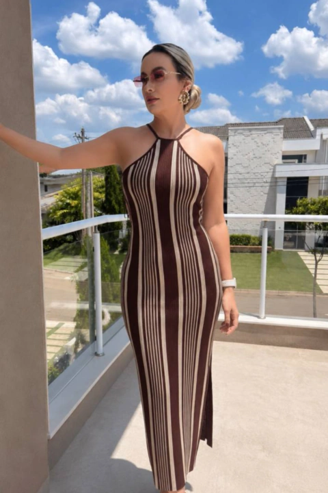 Mulher usando vestido midi justo com listras verticais em tons de marrom e bege, com decote halter, em ambiente externo com céu azul ao fundo