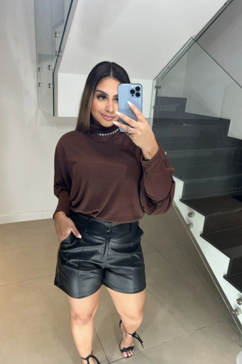 Mulher tirando selfie em espelho vestindo blusa morcego marrom com gola alta e shorts pretos de couro
